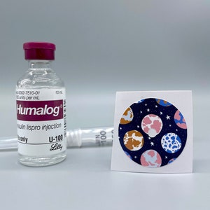 May include: A clear Humalog insulin vial with a red cap, a syringe, and a circular sticker featuring a planet pattern in pink, blue, and brown on a navy background. The vial reads "Humalog" and "insulin lispro injection."