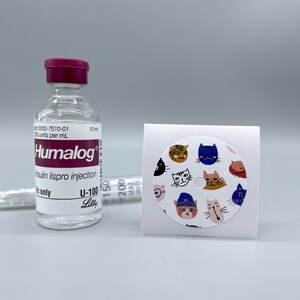 Może przedstawiać: Przezroczysta butelka szklana z fioletowym korkiem z etykietą "Humalog" insulina lispro iniekcja U-100 Lilly. Butelka znajduje się obok białej naklejki z okrągłą, kolorową naklejką przedstawiającą wzór z kocimi twarzami.