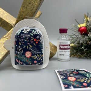 Può includere: Un pod per pompa per insulina bianco con un design natalizio festivo che presenta rami di pino, bacche e fiocchi di neve su uno sfondo blu scuro. Il pod è attaccato a un supporto in metallo dorato. Una piccola bottiglia di insulina Humalog è visibile sullo sfondo.