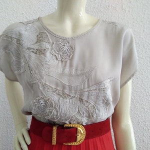 May include: A light gray blouse with intricate floral embroidery and lace detailing. The short-sleeved top features a round neckline and is paired with a red belt with a gold buckle. The blouse is displayed on a mannequin.