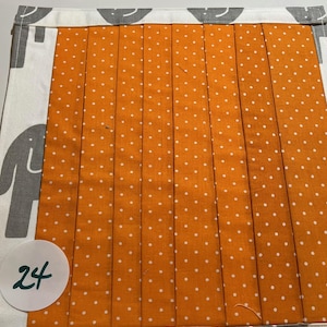 May include: A square, handmade fabric placemat. The center is orange with white polka dots and vertical pleats. The border is white with gray elephant silhouettes and a gray and white checkered trim. A white circle with the number "24" is in the lower left corner.