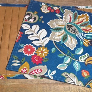 Puede incluir: Dos manteles individuales con diseño floral. Los manteles individuales tienen un fondo azul con flores grandes en rojo, blanco y beige. Un mantel individual está doblado sobre el otro, revelando un interior beige. Una pegatina blanca con el número 138 y la palabra "UPRIGHT" es visible.