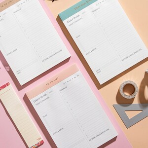 May include: Three daily planner notepad sets with different coloured covers. Each notepad has a daily plan section with today's priorities, to do list, notes and ideas, and a schedule section. The notepads are on a pink surface.