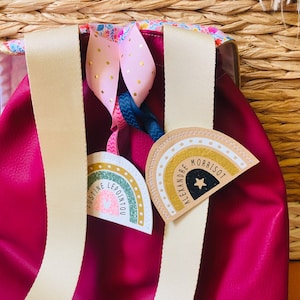 May include: A fuchsia backpack with cream-colored straps and decorative ribbons. Two rainbow-shaped tags are attached, one with the name "JUSTINE LEPOINTOU" and the other with "ALEXANDRE MORRISOT". The tags feature a rainbow design with gold accents.