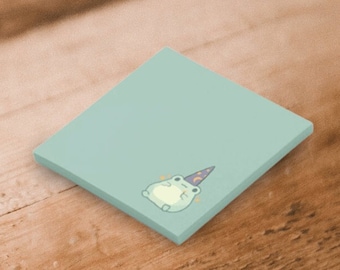 May include: A square light green sticky note featuring a small illustration of a light green frog wearing a purple wizard hat with a yellow crescent moon and star pattern. The frog is positioned in the bottom corner of the note.