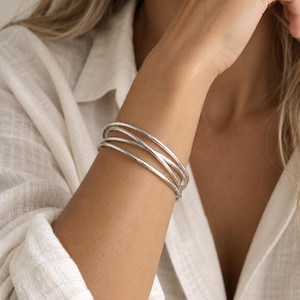 Puede incluir: Una pulsera de tipo brazalete plateado que consta de cuatro bandas circulares finas e interconectadas con un acabado mate y texturizado. Las bandas de metal se superponen y se cruzan entre sí, creando una apariencia de capas en la muñeca.
