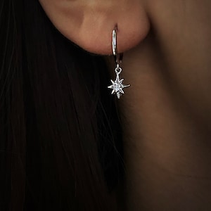 Puede incluir: Pendientes de aro de plata con un colgante de estrella que presenta un cristal transparente en el centro.