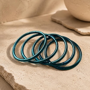 May include: A set of five thin, circular bangles in a deep metallic teal colour. Each bangle features a smooth, reflective surface with a consistent rounded profile. The bracelets are arranged in a slightly overlapping, stacked formation.