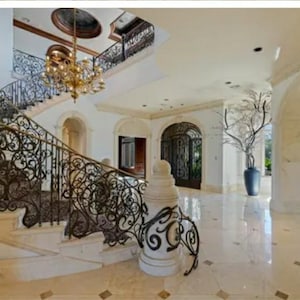 May include: A grand staircase with an ornate wrought iron railing leads up to a landing with a large arched doorway. The staircase is made of white marble and the railing is black.