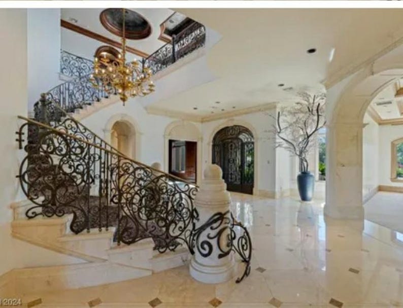 May include: A grand staircase with an ornate wrought iron railing leads up to a landing with a large arched doorway. The staircase is made of white marble and the railing is black.