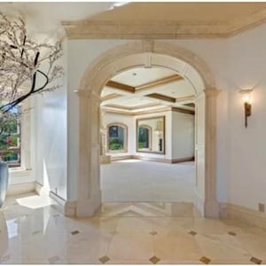 May include: A large, white, arched doorway with ornate trim leads into a spacious room with a tiled floor. A large, dark gray vase with a leafy plant sits in the foreground.