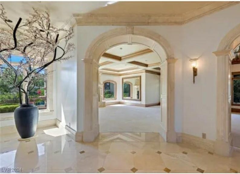 May include: A large, white, arched doorway with ornate trim leads into a spacious room with a tiled floor. A large, dark gray vase with a leafy plant sits in the foreground.