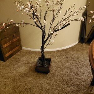 May include: A white faux cherry blossom tree with brown branches in a black planter pot. The tree is on a brown carpet.