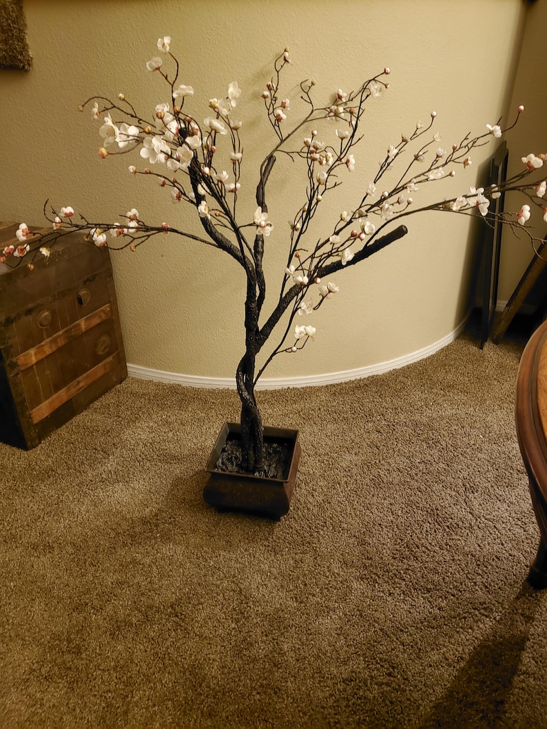 May include: A white faux cherry blossom tree with brown branches in a black planter pot. The tree is on a brown carpet.