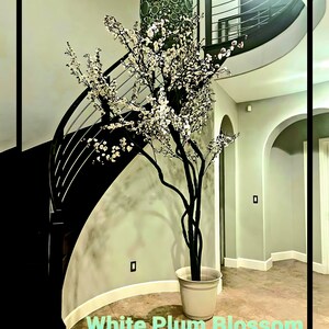 May include: A white artificial plum blossom tree in a white planter pot, standing in a foyer with a curved staircase in the background.