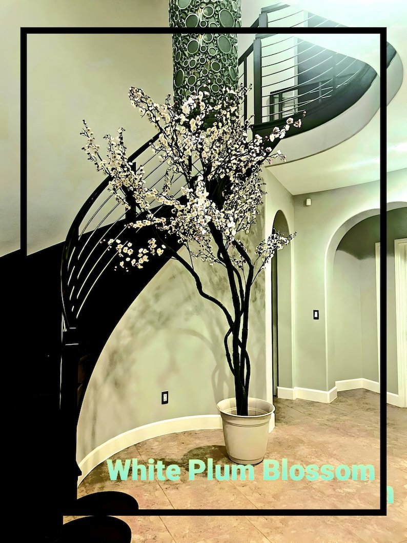 May include: A white artificial plum blossom tree in a white planter pot, standing in a foyer with a curved staircase in the background.