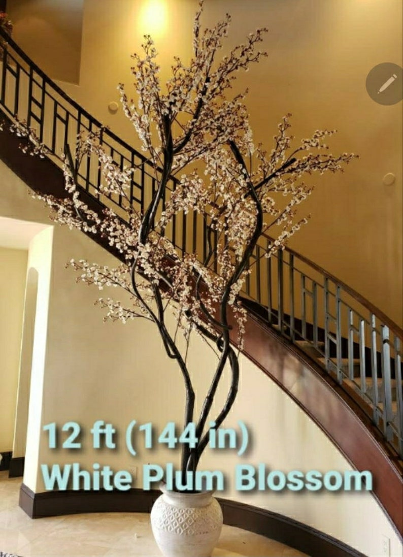 May include: A 12 ft (144 in) tall white plum blossom artificial tree in a white ceramic pot. The tree is displayed in a home setting with a staircase in the background.