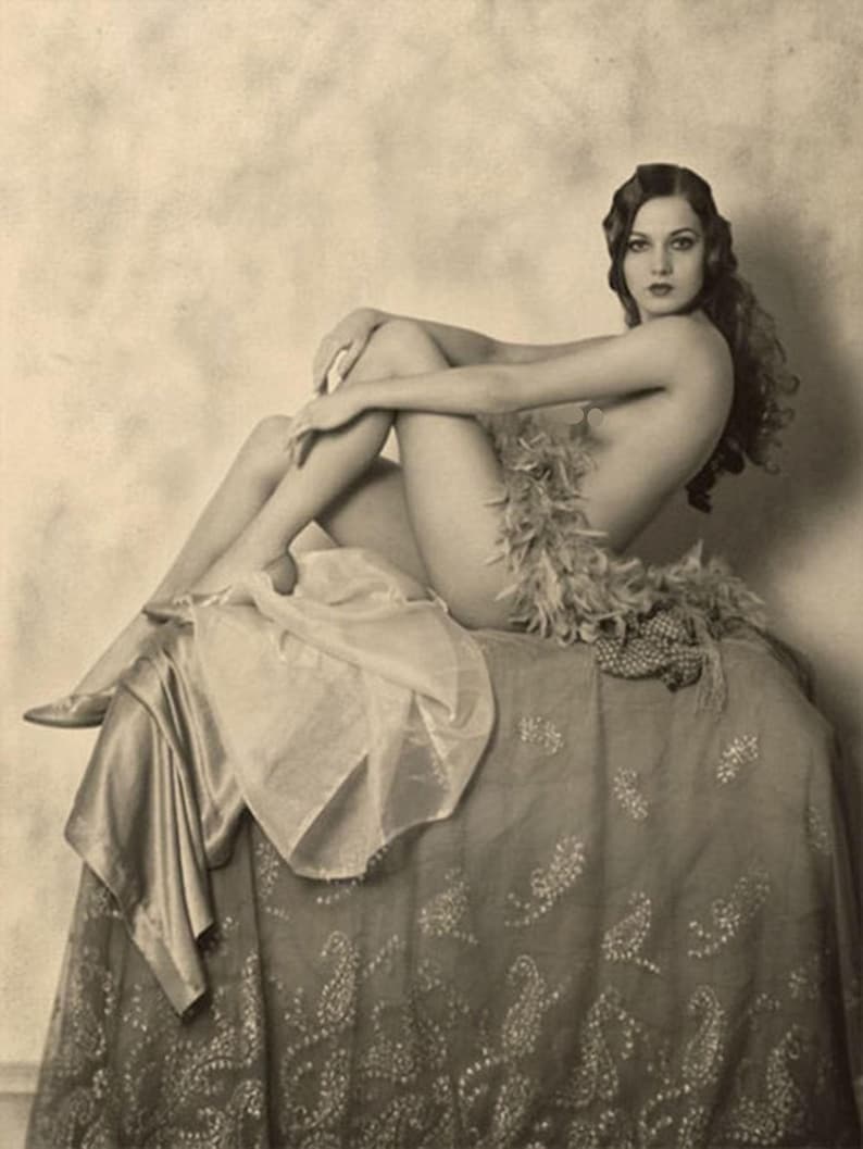 Puede incluir: Una fotograf&iacute;a vintage en blanco y negro de una mujer con un boa de plumas sentada en un taburete cubierto de tela estampada.