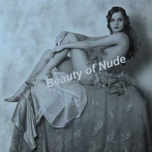Puede incluir: Una fotograf&iacute;a en blanco y negro de una mujer sentada en una cama con un boa de plumas drapeado alrededor de ella. La mujer est&aacute; en una pose simple con las piernas cruzadas y los brazos descansando sobre sus piernas. La cama est&aacute; cubierta con una tela estampada.