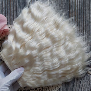 May include: A bundle of wavy, off-white doll hair is held by a gloved hand. A pink rose and a lace doily are next to the hair on a gray wooden surface. The hair has a natural, textured appearance, suitable for doll making.