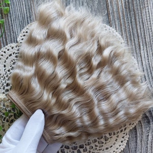 May include: A light blonde wavy hair weft for doll making. The weft is held in a gloved hand and is on a white lace doily.