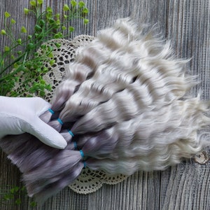 Puede incluir: Un paquete de extensiones de cabello ondulado plateado y gris. El cabello está atado con gomas elásticas azules.