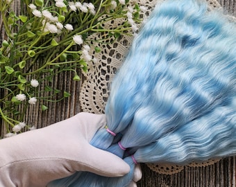 Puede incluir: Un paquete de pelo de muñeca ondulado azul claro, atado con gomas elásticas rosas. El pelo está dispuesto en forma de abanico y está sobre una servilleta blanca.
