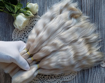 May include: A bundle of platinum blonde human hair wefts, tied together with yellow elastic bands. The wefts are arranged in a fan shape and are lying on a white lace doily.