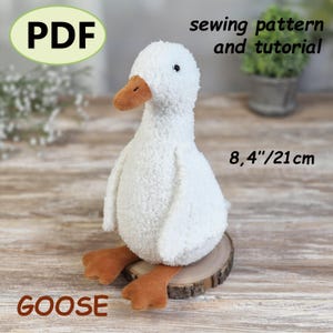 May include: A white plush goose toy with a textured fabric body, orange beak, and orange webbed feet. The image includes the text PDF, sewing pattern and tutorial, 8.4 inches, and GOOSE. The goose stands 8.4 inches tall as indicated by the text.