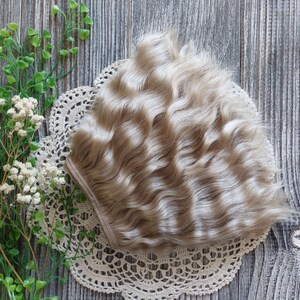 Puede incluir: Una peluca ondulada de color rubio claro para mu&ntilde;eca. La peluca est&aacute; sobre un encaje de encaje blanco sobre una superficie de madera.