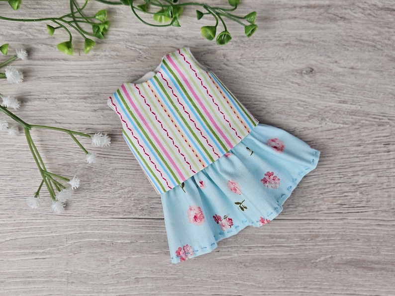 Completed doll outfit with colorful top and floral blue skirt displayed flat with decorative greenery