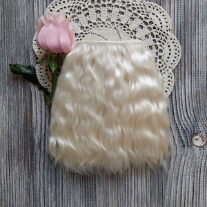 May include: A bundle of wavy, light blonde hair extensions. The hair is arranged on a gray wooden surface, next to a pink rose and a cream-colored lace doily. The hair has a natural, flowing appearance.