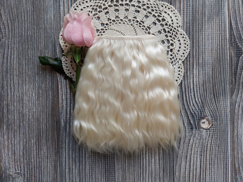 May include: A bundle of wavy, light blonde hair extensions. The hair is arranged on a gray wooden surface, next to a pink rose and a cream-colored lace doily. The hair has a natural, flowing appearance.