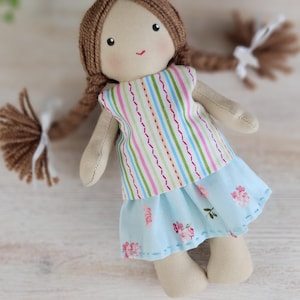Cloth doll dressed in striped top and floral blue skirt standing on wooden surface, handmade toy clothes