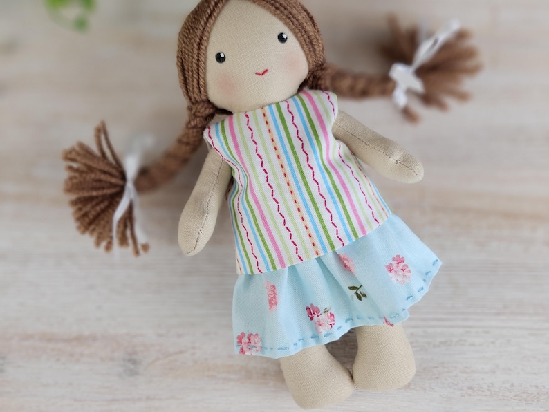 Cloth doll dressed in striped top and floral blue skirt standing on wooden surface, handmade toy clothes