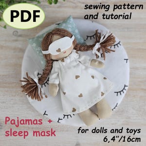 May include: A handmade doll wearing a white nightgown with heart-shaped accents and a sleep mask. The doll is lying on a round pillow with a printed eyelash design. The image includes the text "PDF", "sewing pattern and tutorial", "Pajamas + sleep mask", and "for dolls and toys 6,4"/16cm".