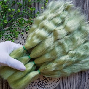 Puede incluir: Un paquete de pelo de muñeca ondulado verde claro, atado con gomas elásticas verdes. El pelo está dispuesto en forma de abanico sobre un encaje blanco.