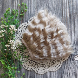 Puede incluir: Una peluca de mu&ntilde;eca rubia clara con cabello ondulado. La peluca est&aacute; sobre un encaje de encaje blanco y un fondo de madera.