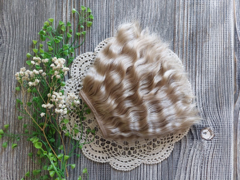 Puede incluir: Una peluca de mu&ntilde;eca rubia clara con cabello ondulado. La peluca est&aacute; sobre un encaje de encaje blanco y un fondo de madera.
