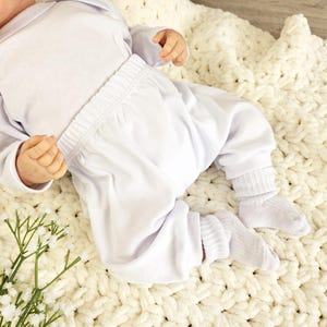 K&ouml;nnte beinhalten: Ein wei&szlig;es Baby-Outfit mit einem lang&auml;rmeligen Shirt und einer Hose. Das Outfit liegt auf einer wei&szlig;en H&auml;keldecke.