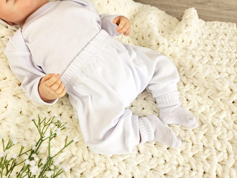 K&ouml;nnte beinhalten: Ein wei&szlig;es Baby-Outfit mit einem lang&auml;rmeligen Shirt und einer Hose. Das Outfit liegt auf einer wei&szlig;en H&auml;keldecke.