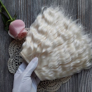 May include: A bundle of wavy, off-white hair extensions. A pink rose and a lace doily are also in the image. The hair is a light, creamy color, and the texture appears soft and voluminous. The background is a wooden surface.