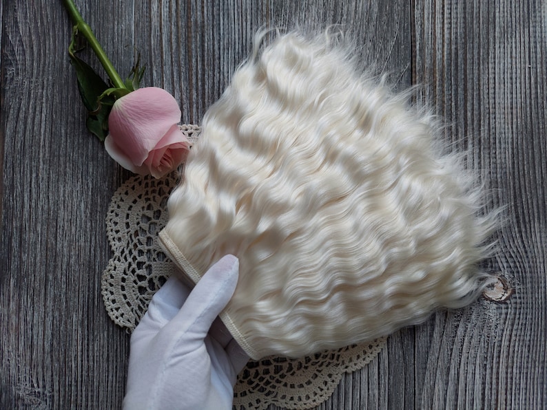 May include: A bundle of wavy, off-white hair extensions. A pink rose and a lace doily are also in the image. The hair is a light, creamy color, and the texture appears soft and voluminous. The background is a wooden surface.