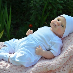 K&ouml;nnte beinhalten: Eine wei&szlig;e Baby Puppe, die einen wei&szlig;en Strampler und eine wei&szlig;e M&uuml;tze tr&auml;gt, liegt auf einer rosa gestrickten Decke.