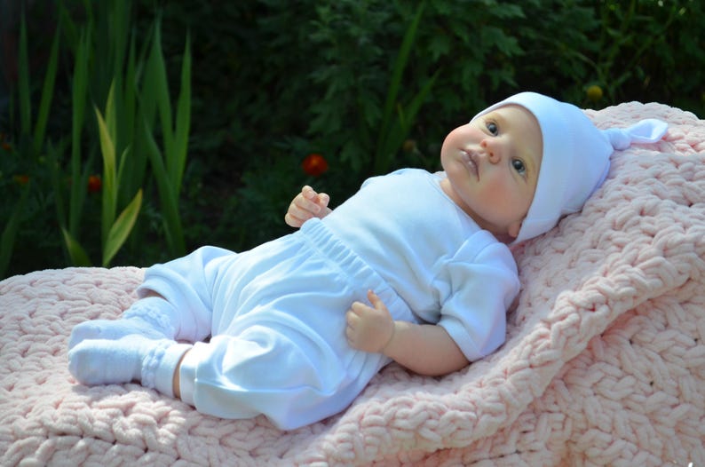 K&ouml;nnte beinhalten: Eine wei&szlig;e Baby Puppe, die einen wei&szlig;en Strampler und eine wei&szlig;e M&uuml;tze tr&auml;gt, liegt auf einer rosa gestrickten Decke.
