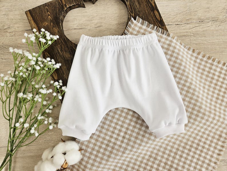 K&ouml;nnte beinhalten: Wei&szlig;e Baby-Bl&uuml;mchen aus Baumwolle mit einem gerafften elastischen Bund. Die Bl&uuml;mchen liegen auf einem braunen und wei&szlig;en karierten Stoff.