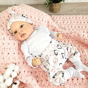 K&ouml;nnte beinhalten: Ein Baby tr&auml;gt einen wei&szlig;en Strampler und eine rosa-schwarze Hose mit Herzmuster. Das Baby tr&auml;gt ein rosa-wei&szlig;es, geknotetes Stirnband mit Herzen darauf. Das Baby liegt auf einer rosa gestrickten Decke.