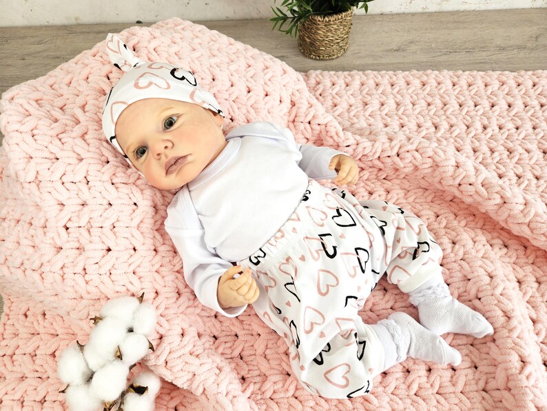 K&ouml;nnte beinhalten: Ein Baby tr&auml;gt einen wei&szlig;en Strampler und eine rosa-schwarze Hose mit Herzmuster. Das Baby tr&auml;gt ein rosa-wei&szlig;es, geknotetes Stirnband mit Herzen darauf. Das Baby liegt auf einer rosa gestrickten Decke.