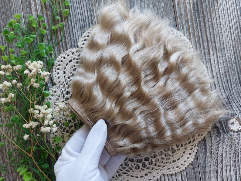 May include: A light blonde wavy hair weft for doll making. The weft is held in a gloved hand and is on a white lace doily.