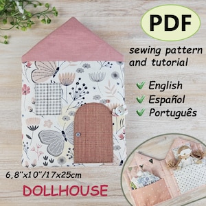 May include: A fabric dollhouse with a pink roof and a floral patterned facade. The dollhouse features a brown door, a window, and a button closure. The text "DOLLHOUSE" is displayed at the bottom. Dimensions are 17cm x 25cm.